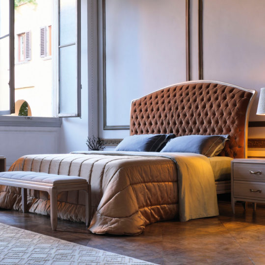 A cozy bedroom features a large Classic Italian Button Upholstered Bed in soft, light brown fabric. The bed is neatly made with a tan comforter and blue pillows. Two wooden nightstands with lamps flank the bed. There is a cushioned bench at the foot of the bed, and large windows in the background.