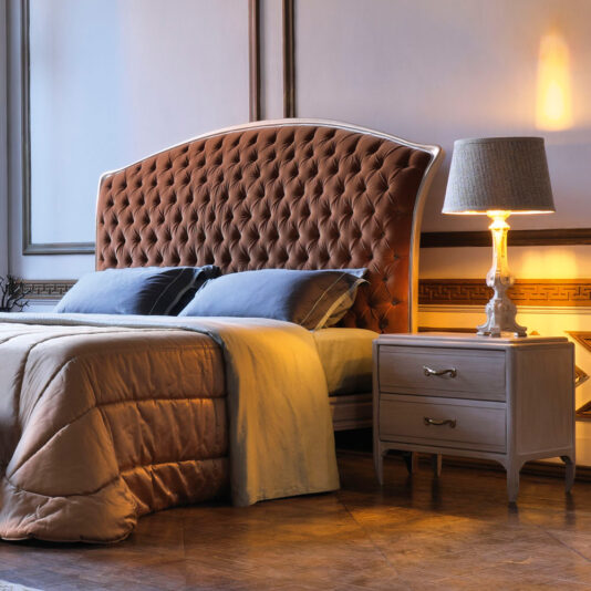 A cozy bedroom features the Classic Italian Button Upholstered Bed with a tufted brown headboard, adorned with gray and beige bedding and a warm-toned throw blanket. To the right of the bed is a wooden nightstand with two drawers, topped by a lamp. The room is softly lit, creating a warm ambiance.