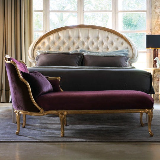 Elegant bedroom with a tufted, white upholstered headboard, dark grey bedding, and large windows in the background. A luxurious Classic Louis XVI Style Chaise Longue with ornate gold trim sits prominently in the foreground on a grey area rug. Beige curtains frame the windows beautifully.