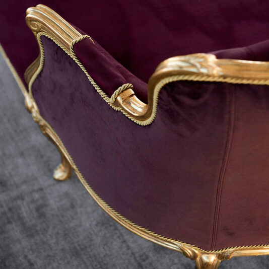 Close-up view of the Classic Louis XVI Style Chaise Longue, elegantly upholstered in rich purple velvet with intricate gold detailing on the edges. The armchair is positioned on a gray textured floor, exuding an opulent and luxurious ambiance.