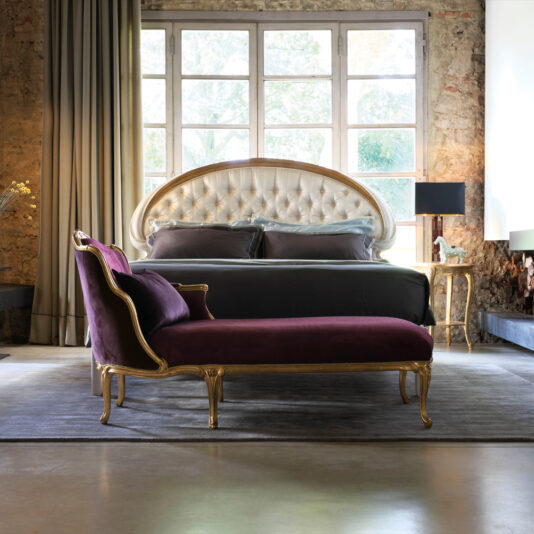 A luxurious bedroom showcases a large tufted headboard bed positioned against a rustic brick wall with expansive windows. At the foot of the bed is the exquisite Classic Louis XVI Style Chaise Longue, adorned in purple velvet and gold trim. The space is elegantly completed with a gray rug, nightstands, lamps, and heavy curtains.