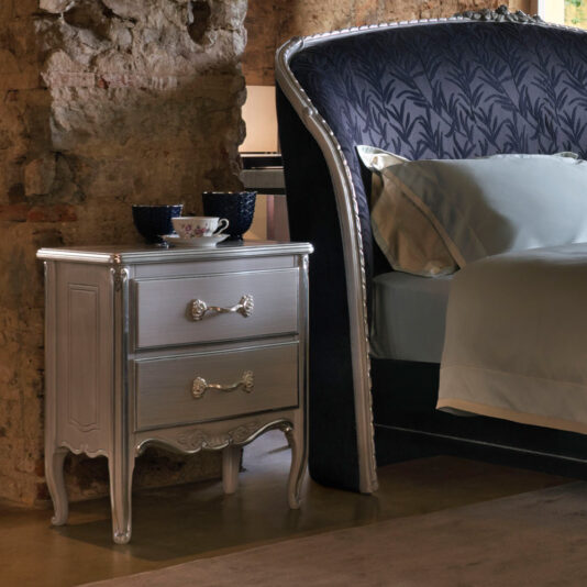 A luxury bedroom scene with a Classic Ornate Bedside Cabinet next to an equally lavish bed. The cabinet features two drawers with decorative handles and is adorned with a teacup, saucer, and two mugs. The wall boasts an exposed, rustic brick texture.