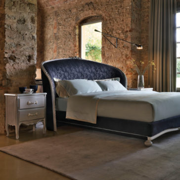 Classic Ornate Bedside Cabinet A cozy bedroom with a stone wall and large window. The bed features an ornate, high, curved headboard with dark patterns and is dressed in light-colored bedding. A Classic Ornate Bedside Cabinet with drawers holds a lamp, vase, and cups. A vintage-style nightstand complements the floor rug to complete the space.