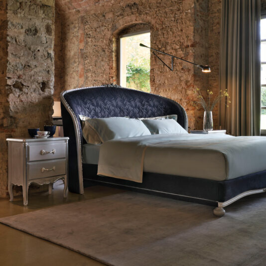 A cozy bedroom with a stone wall and large window. The bed features an ornate, high, curved headboard with dark patterns and is dressed in light-colored bedding. A Classic Ornate Bedside Cabinet with drawers holds a lamp, vase, and cups. A vintage-style nightstand complements the floor rug to complete the space.