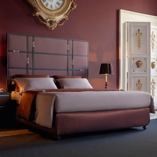 A luxurious bedroom with dark red walls, showcasing a Classic Snaffle Upholstered Bed With Tall Headboard dressed in maroon and gray bedding. An ornate gold clock is mounted above the bed, while a side table with a lamp sits adjacent. The room also features a white door embellished with gold accents.