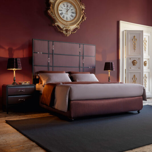 Classic Snaffle Upholstered Bed With Tall Headboard A luxurious bedroom showcases a Classic Snaffle Upholstered Bed With Tall Headboard, accompanied by two bedside tables with matching lamps. The room is enhanced by rich, deep red walls, a vintage clock above the headboard, and an ornate white door with gold accents.