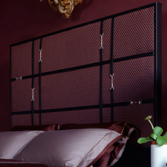 Classic Snaffle Upholstered Bed With Tall Headboard A modern bedroom features the Classic Snaffle Upholstered Bed With Tall Headboard, adorned with distinctive hooks. The wall behind the bed is painted a rich burgundy, while the bedding includes pillows and sheets in various shades of brown. A potted plant on a nearby surface adds a touch of nature to the space.