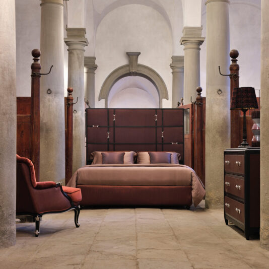 Classic Snaffle Upholstered Bed With Tall Headboard A stylish bedroom features the Classic Snaffle Upholstered Bed With Tall Headboard, paired with matching pillows and duvet, set against tall, arched columns. To the left sits a maroon armchair, while to the right stands a dark wooden dresser topped with a decorative lamp. The room is accented by a rustic stone floor.