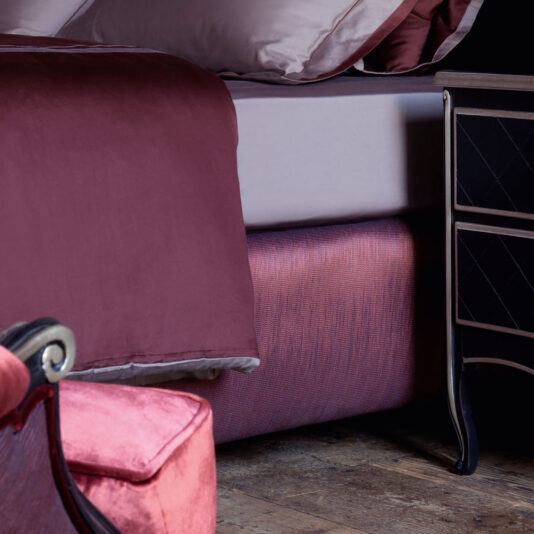 Classic Snaffle Upholstered Bed With Tall Headboard A cozy bedroom showcases the Classic Snaffle Upholstered Bed With Tall Headboard, adorned with mauve sheets. Beside the bed stands a black nightstand featuring a diamond pattern, while a plush pink armchair is partially visible in the foreground.