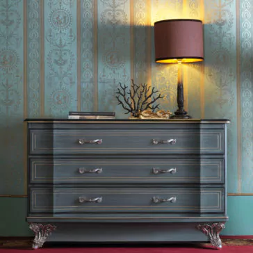 Classic Style Italian Chest Of Drawers A Classic Style Italian Chest Of Drawers with ornate silver handles and claw feet is positioned against a wallpapered wall with intricate patterns. A table lamp with a brown shade, a decorative branch sculpture, and two books rest on top of the chest.