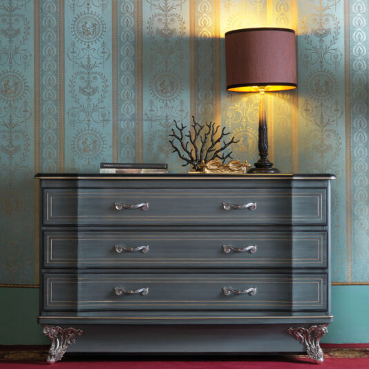 A Classic Style Italian Chest Of Drawers with ornate silver handles and claw feet is positioned against a wallpapered wall with intricate patterns. A table lamp with a brown shade, a decorative branch sculpture, and two books rest on top of the chest.