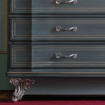 Classic Style Italian Chest Of Drawers A close-up of the Classic Style Italian Chest Of Drawers showcases its dark, elegant design with three compartments featuring silver handles and intricate gold trim. The chest is adorned with a detailed, silver decorative claw foot at the bottom, resting on red carpeting.
