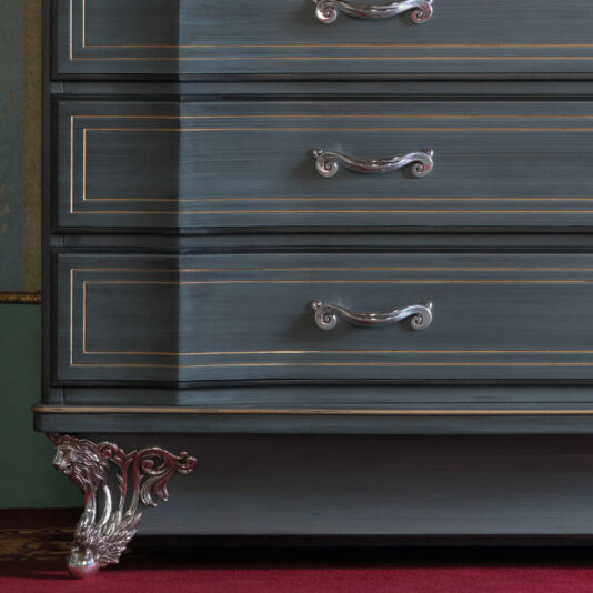 A close-up of the Classic Style Italian Chest Of Drawers showcases its dark, elegant design with three compartments featuring silver handles and intricate gold trim. The chest is adorned with a detailed, silver decorative claw foot at the bottom, resting on red carpeting.
