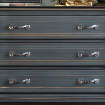 Classic Style Italian Chest Of Drawers A close-up of the Classic Style Italian Chest Of Drawers showcasing its three drawers, each adorned with two ornate, silver handles featuring a swirl design. The dresser boasts a blue-grey finish with subtle gold accents, exuding a timeless elegance. The top surface is adorned with various items such as books and decorative objects.
