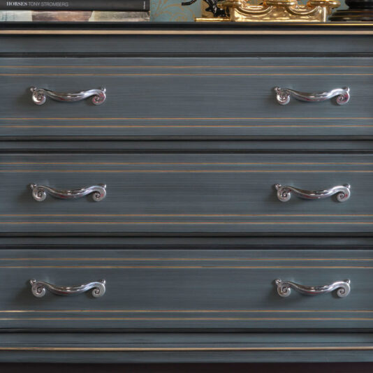 A close-up of the Classic Style Italian Chest Of Drawers showcasing its three drawers, each adorned with two ornate, silver handles featuring a swirl design. The dresser boasts a blue-grey finish with subtle gold accents, exuding a timeless elegance. The top surface is adorned with various items such as books and decorative objects.