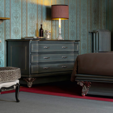 Classic Style Italian Chest Of Drawers A vintage-style bedroom with patterned wallpaper showcases a Classic Style Italian Chest Of Drawers, elegantly designed with ornate metal legs. This dresser, decorated with books, a lamp with a shade, and various decorative items, complements the matching dark wood bed frame partially visible under a brown blanket.