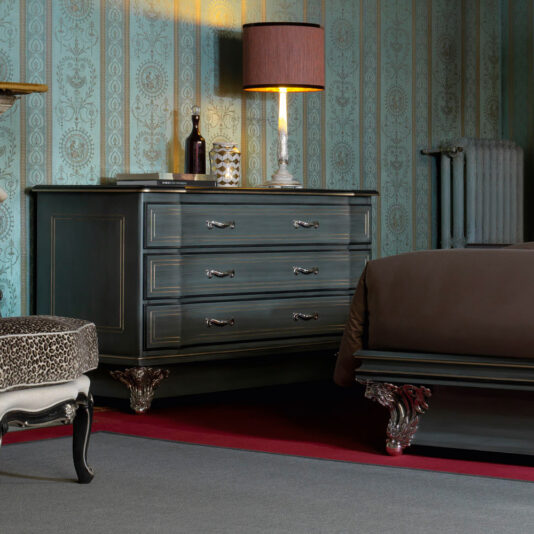 A vintage-style bedroom with patterned wallpaper showcases a Classic Style Italian Chest Of Drawers, elegantly designed with ornate metal legs. This dresser, decorated with books, a lamp with a shade, and various decorative items, complements the matching dark wood bed frame partially visible under a brown blanket.