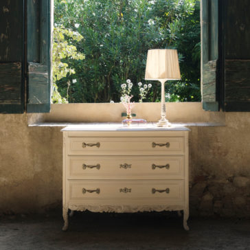 Classic Style Ornate Chest Of Drawers A Classic Style Ornate Chest Of Drawers with intricate handles and carvings sits near an open window with rustic wooden shutters. A delicate table lamp with a pleated shade and a small glass vase with flowers rest on the high-end modern chest of drawers. Lush greenery is visible outside the window.