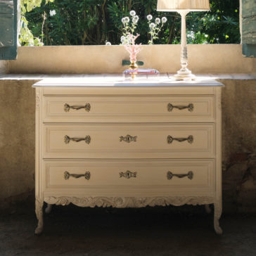 Classic Style Ornate Chest Of Drawers The Classic Style Ornate Chest Of Drawers is a vintage white wooden dresser with intricate carvings, featuring three drawers adorned with ornate brass handles. It seamlessly blends as a high-end modern chest of drawers, standing against a sunlit wall with an open window above, framed by green shutters. Atop it sits a lamp, glass figurine, and small box.