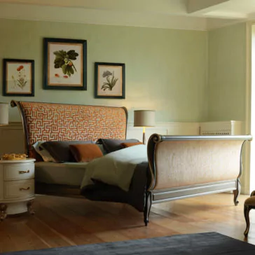 Classic Style Upholstered Sleigh Bed A luxurious bedroom showcases the Classic Style Upholstered Sleigh Bed, featuring an ornate frame and a patterned headboard and footboard. The bed is adorned with pillows and bedding in muted tones. Two framed botanical prints hang on a light green wall, while two side tables with lamps flank the bed.