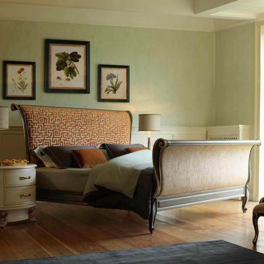 A luxurious bedroom showcases the Classic Style Upholstered Sleigh Bed, featuring an ornate frame and a patterned headboard and footboard. The bed is adorned with pillows and bedding in muted tones. Two framed botanical prints hang on a light green wall, while two side tables with lamps flank the bed.