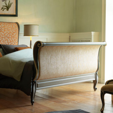 Classic Style Upholstered Sleigh Bed A stylish bedroom showcases the Classic Style Upholstered Sleigh Bed, with its elegant beige headboard and footboard. The bed is dressed in neutral-toned pillows and bedding. On the bedside table, a lamp with a patterned shade illuminates the space as sunlight pours in through a nearby window.