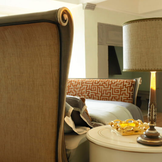 A close-up view of a bedroom featuring the Classic Style Upholstered Sleigh Bed, showcasing its elegant design with rolled tops on the headboard and footboard. To the right, a round bedside table holds a stylish lamp with a woven shade and a gold decorative dish. The bed is partially seen with patterned pillows.