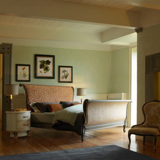 A cozy bedroom with a large, Classic Style Upholstered Sleigh Bed featuring an intricately patterned headboard and footboard. The room has hardwood floors, light-colored walls, framed botanical prints above the bed, a round bedside table, two lamps, and a cushioned chair. Light filters through a window.