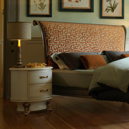 A cozy bedroom showcases the Classic Style Upholstered Sleigh Bed, highlighted by its ornate orange and white patterned headboard. The bed is adorned with brown and green pillows for added comfort. Beside the bed sits a white nightstand with gold hardware, topped with a lamp. Wooden flooring and framed botanical prints further enhance the room's warm ambiance.
