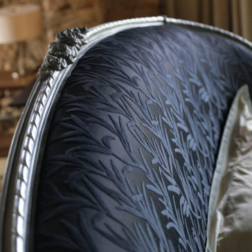 Classic Style Upholstered Winged Bed A close-up showcases the ornate headboard of the Classic Style Upholstered Winged Bed, featuring dark-colored, textured upholstery with a detailed woven floral pattern. The headboard is adorned with a carved metallic frame complete with decorative flourishes. In the background, a muted-tone interior is softly blurred.