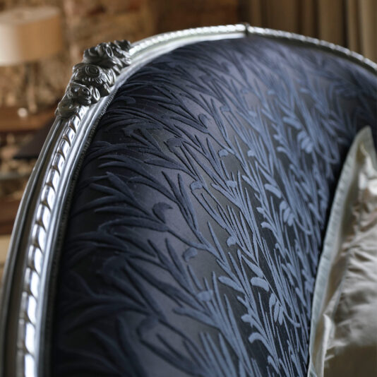 A close-up showcases the ornate headboard of the Classic Style Upholstered Winged Bed, featuring dark-colored, textured upholstery with a detailed woven floral pattern. The headboard is adorned with a carved metallic frame complete with decorative flourishes. In the background, a muted-tone interior is softly blurred.