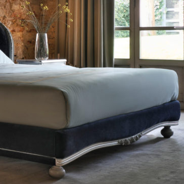 Classic Style Upholstered Winged Bed A cozy bedroom features the Classic Style Upholstered Winged Bed, boasting a plush navy-blue frame and light gray bedding. A side table holding a vase of thin branches and buds sits beside the bed. The room is adorned with a rustic stone wall and large windows with curtains, allowing natural light to fill the space.