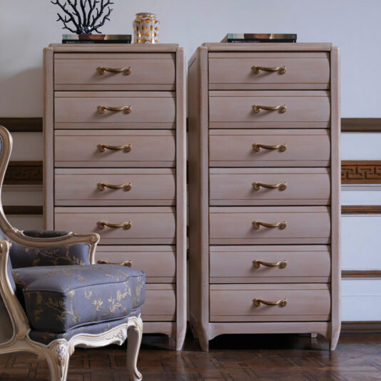 A pair of Classic Tallboy Chest Of Drawers with gold handles stand against a wall adorned with decorative trim. In front of them, a blue upholstered chair featuring a floral pattern adds charm to the room, which boasts parquet flooring. Decorative items sit atop the dressers.