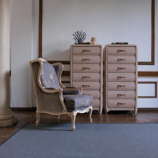 An elegant room features a cushioned armchair with ornate wooden trim and floral upholstery. Flanking the chair are two matching tall, light-toned Classic Tallboy Chests of Drawers with gold handles, set against a white wall accented in brown. A large gray rug covers the wooden floor.
