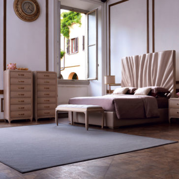 A luxurious bedroom with elegant furnishings features a bed with a tall, tufted headboard, a soft pink duvet, nightstands, the Classic Tallboy Chest Of Drawers for ample storage, and an upholstered bench at the foot of the bed. The room is accentuated by high ceilings and a large window.