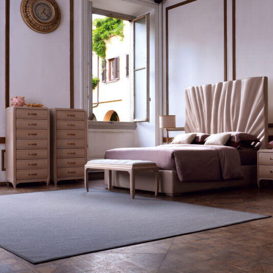 A luxurious bedroom with elegant furnishings features a bed with a tall, tufted headboard, a soft pink duvet, nightstands, the Classic Tallboy Chest Of Drawers for ample storage, and an upholstered bench at the foot of the bed. The room is accentuated by high ceilings and a large window.