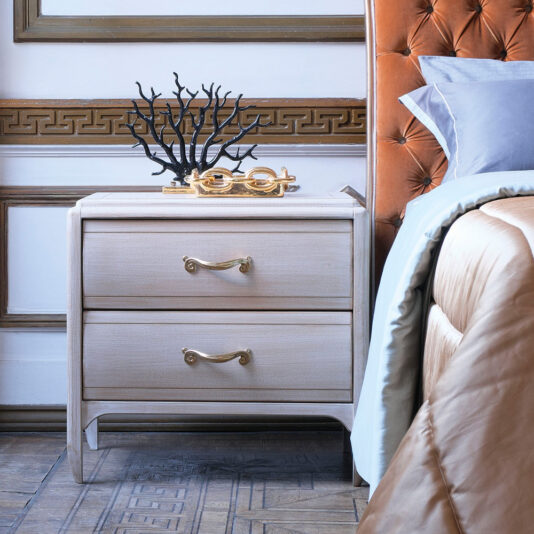 A neatly arranged bedroom features a Classic Two Drawer Bedside Cabinet with brass handles. Atop the nightstand are a black coral sculpture and a golden figurine. To the right, part of a bed with a tufted leather headboard and blue bedding can be seen.