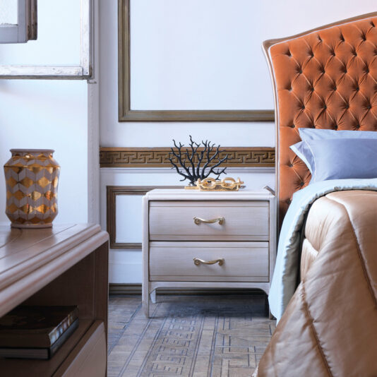 Classic Two Drawer Bedside Cabinet A stylish bedroom featuring a tufted orange headboard, a **Classic Two Drawer Bedside Cabinet** in beige with intricate detailing, and a decorative black coral ornament. The room boasts elaborate wall molding and a patterned wooden floor. A geometric vase sits gracefully on a nearby console table.