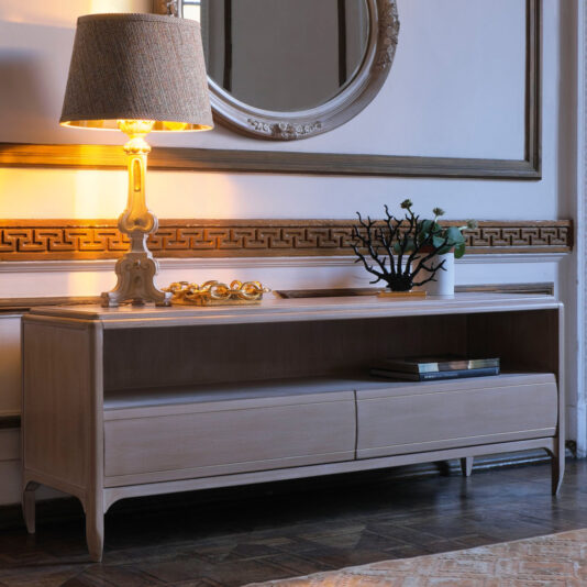 The Classic Two Drawer TV Unit, which also serves as a wooden console with an open shelf, is enhanced with a decorative lamp, a round mirror above it, and a minimalist plant sculpture. This setup is placed against an elegant wall featuring intricate designs and wooden trim, establishing a sophisticated ambiance.