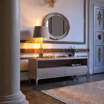 Classic Two Drawer TV Unit A vintage-style living room corner features the Classic Two Drawer TV Unit, adorned with a decorative table lamp, a floral arrangement, and other small decor items. Above the unit hangs a large, ornate round mirror. The room boasts a classic design with intricate molding and a polished parquet floor.