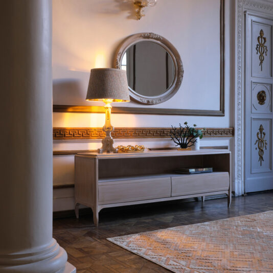 Classic Two Drawer TV Unit A vintage-style living room corner features the Classic Two Drawer TV Unit, adorned with a decorative table lamp, a floral arrangement, and other small decor items. Above the unit hangs a large, ornate round mirror. The room boasts a classic design with intricate molding and a polished parquet floor.
