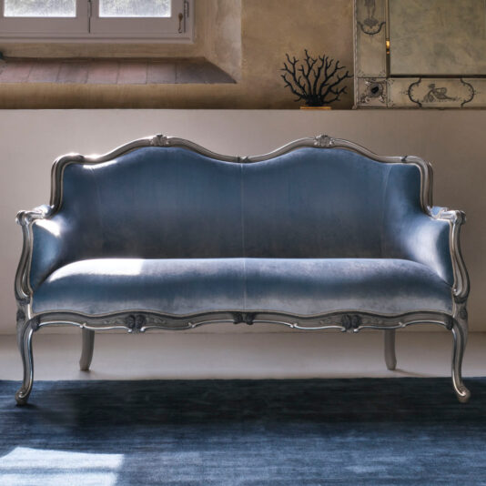 The Classic Venetian Style 2 Seater Sofa, upholstered in blue velvet and featuring an intricately carved silver frame, sits against a beige wall. The antique-style sofa is positioned on a dark blue rug near a window, reflecting a blend of elegance and classic design.