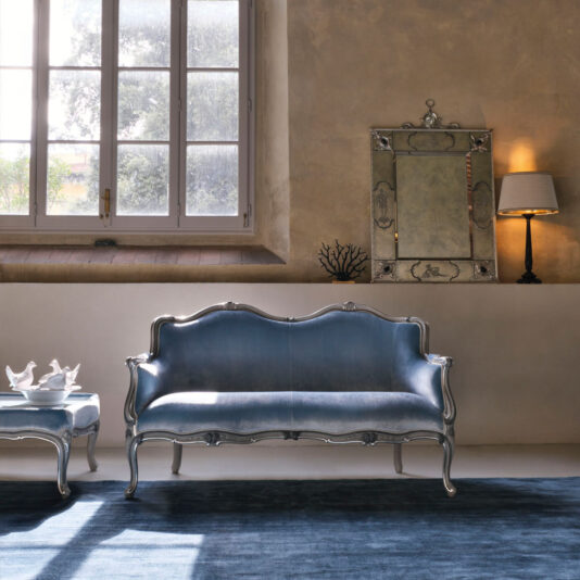 A stylish living room showcases a vintage blue Classic Venetian Style 2 Seater Sofa accompanied by a matching armchair. Natural light streams through a large window, illuminating the blue rug below. Above the sofa, an ornate mirror enhances the beige wall, complemented by a nearby wall-mounted lamp. A white teapot set rests on the chair.