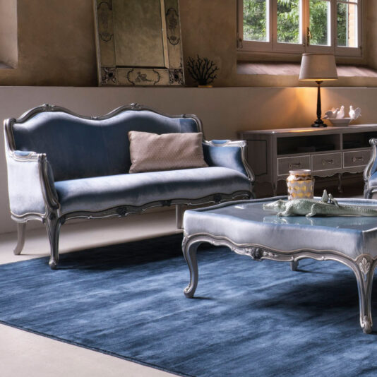 A chic living room boasts an ornate Classic Venetian Style 2 Seater Sofa with blue cushions and matching ottoman, both featuring elegant silver frames. The floor is adorned with a navy blue rug, while a white dresser adorned with a lamp, decor items, and a framed mirror sits in the background. Sunlight pours in through the window, casting a warm glow across the space.