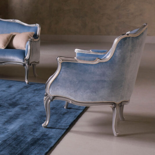 A sophisticated living room scene featuring elegant furniture: the Classic Venetian Style Armchair in blue velvet with metallic accents and curved legs, paired with a matching blue velvet loveseat in the background. The interior decor includes a soft blue area rug on a polished floor.