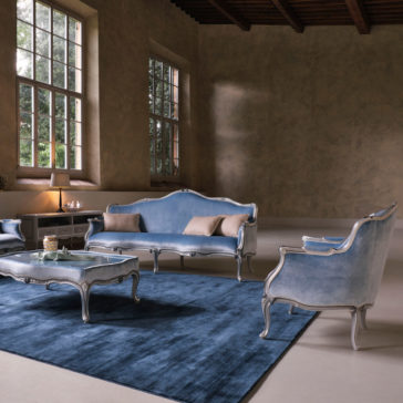 Classic Venetian Style Armchair A stylish living room features blue velvet antique-style furniture, including a sofa, the Classic Venetian Style Armchair, and a coffee table, all set on a large blue rug. Tall windows on the beige wall allow natural light to fill the spacious room. A lit table lamp is on one side.