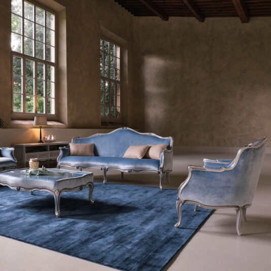Classic Venetian Style Armchair A stylish living room features blue velvet antique-style furniture, including a sofa, the Classic Venetian Style Armchair, and a coffee table, all set on a large blue rug. Tall windows on the beige wall allow natural light to fill the spacious room. A lit table lamp is on one side.