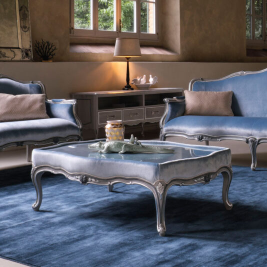 A stylish, plush living room features vintage blue seating with ornate silver trim, including a classic Venetian style armchair. A Classic Venetian Style Upholstered Coffee Table adorned with a decorative alligator centerpiece and a blue rug complete the scene. A lamp emits soft lighting near a window with trees visible outside.