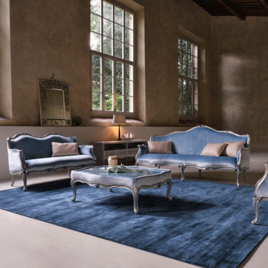 Classic Venetian Style Upholstered Coffee Table A spacious room with large windows features elegant vintage furniture, including a blue velvet sofa set with a matching loveseat and a classic Venetian style armchair. The centerpiece is the Classic Venetian Style Upholstered Coffee Table that sits on a dark blue area rug. A lamp, mirror, and some decor items complete the setting.