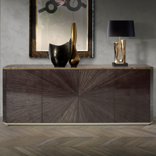The Contemporary Grey Buffet, featuring doors with a striking sunburst pattern, is positioned against a textured gray wall. Its top is adorned with abstract sculptures, a gold-framed painting, and a black lamp with a gold base. The floor showcases chevron-patterned wooden planks.